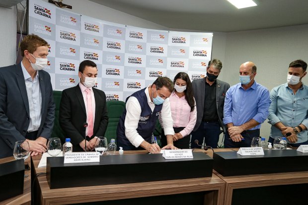 Governador Carlos Moisés assinando documento em mesa de reunião com seis autoridades, formalizando a ampliação de 30 leitos de UTI para hospitais do Extremo Sul catarinense.