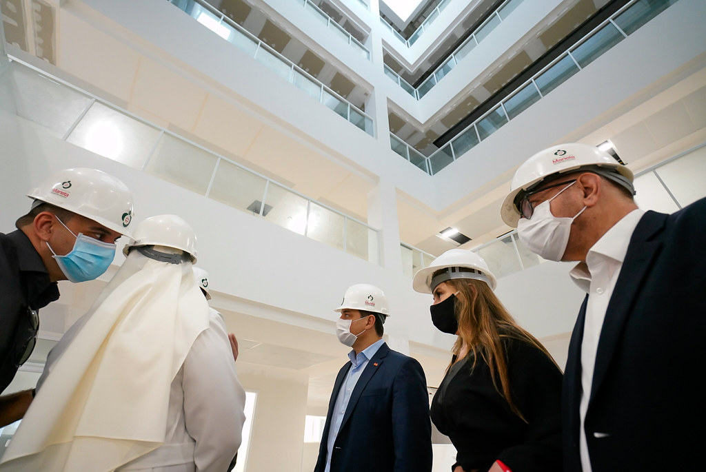 Grupo de pessoas usando capacetes de proteção e máscaras faciais visita o interior de um prédio moderno, com vários andares e varandas internas visíveis ao fundo. Entre os visitantes há homens e uma mulher, todos vestidos formalmente, aparentando participar de uma inspeção ou visita técnica à construção.