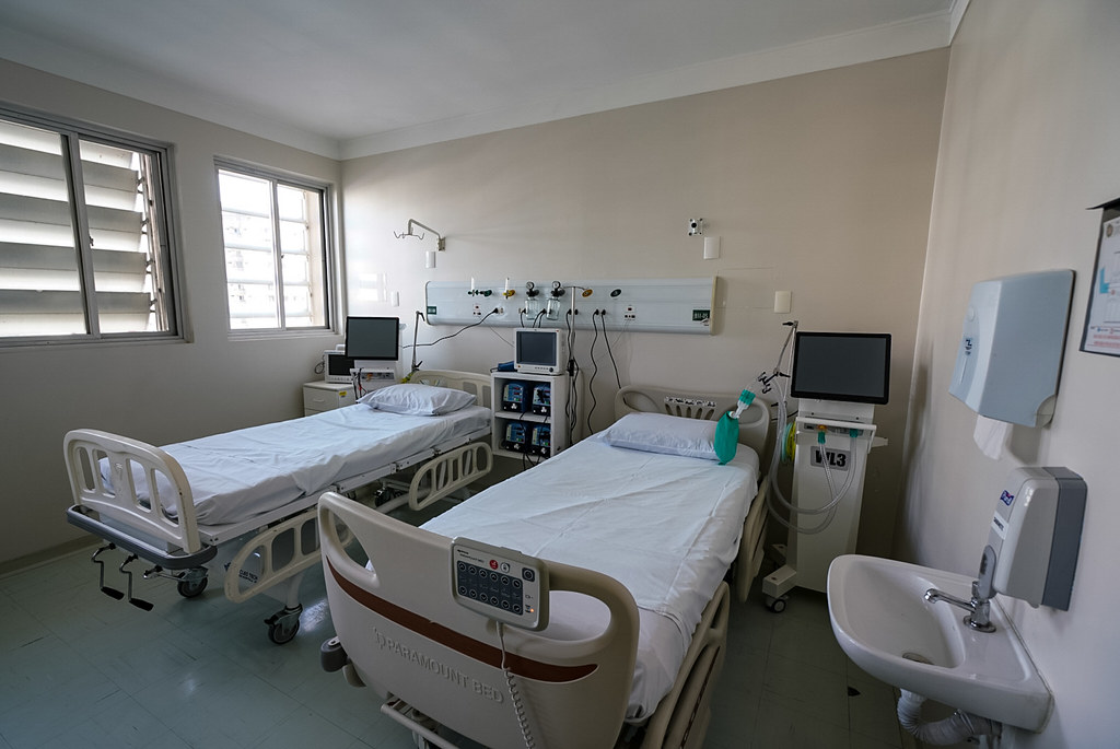 Quarto hospitalar com duas camas equipadas com lençóis brancos, monitores e aparelhos médicos ao lado de cada leito. O ambiente é limpo, iluminado por janelas grandes, e há uma pia com dispensador de sabonete fixado na parede.