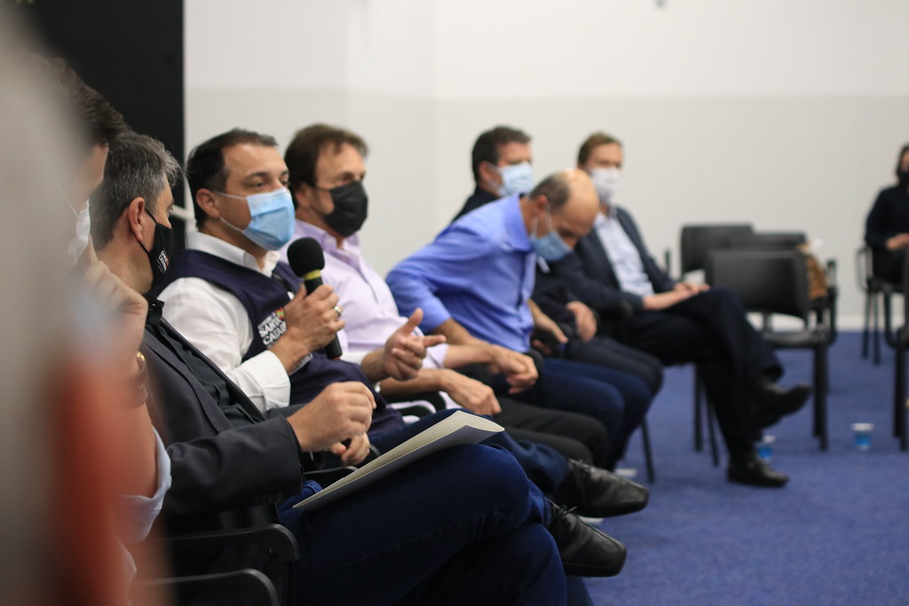 Governador Carlos Moisés de traje formal e máscara discursando em microfone, acompanhado por autoridades sentadas ao seu lado, durante anúncio de medidas no Extremo Sul catarinense.