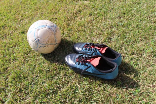 Bola de futebol branca com detalhes azuis e um par de chuteiras pretas e azuis com cadarços pretos, posicionados sobre a grama verde sob luz do sol.