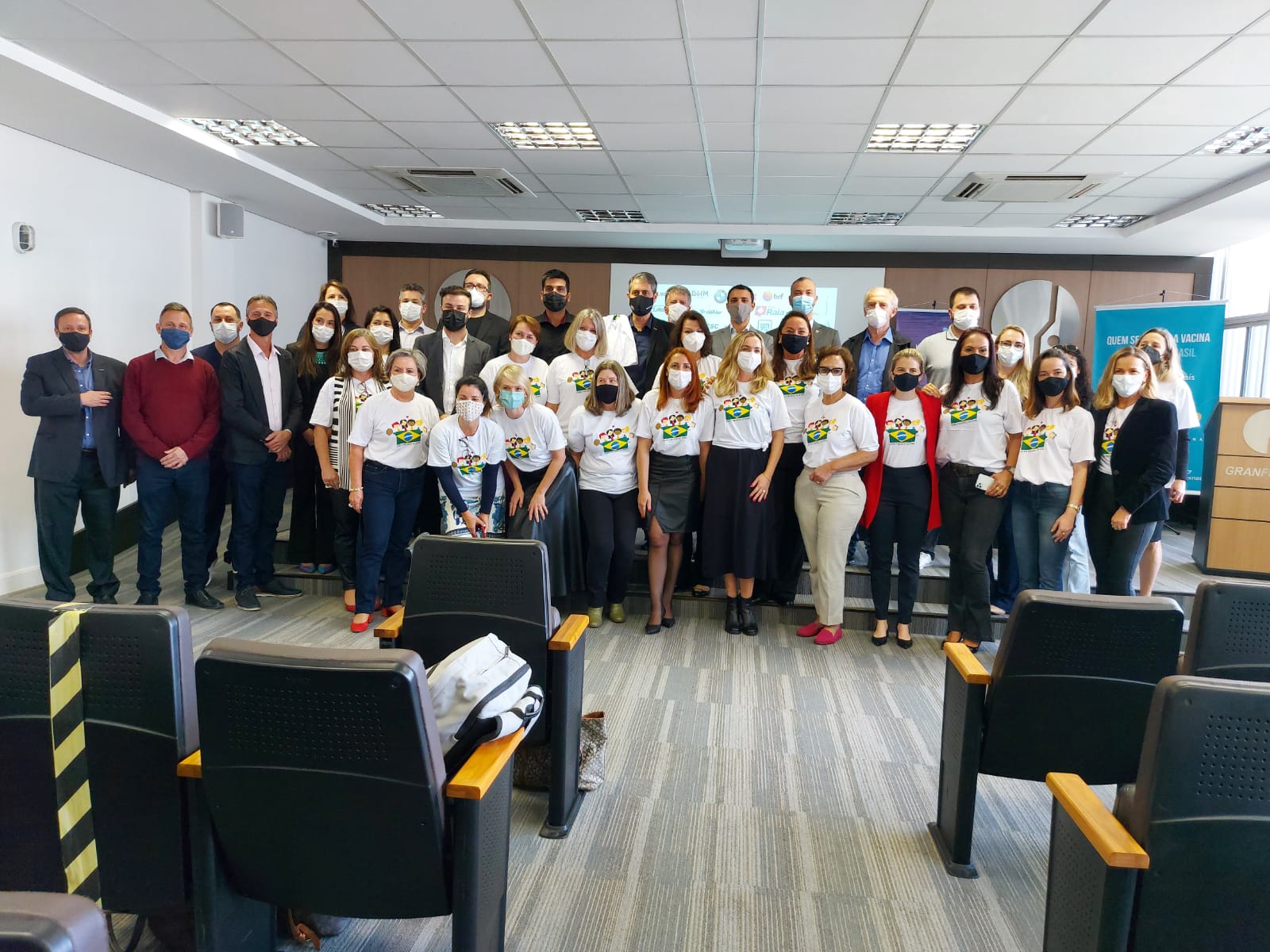 Grupo grande de pessoas posando para foto em uma sala de conferências. Muitos usam camisetas brancas com estampa colorida e todos estão de máscara. Ao fundo, banners institucionais e o logotipo da GRANPOLIS na parede. O ambiente é iluminado e formal, típico de evento ou reunião oficial.