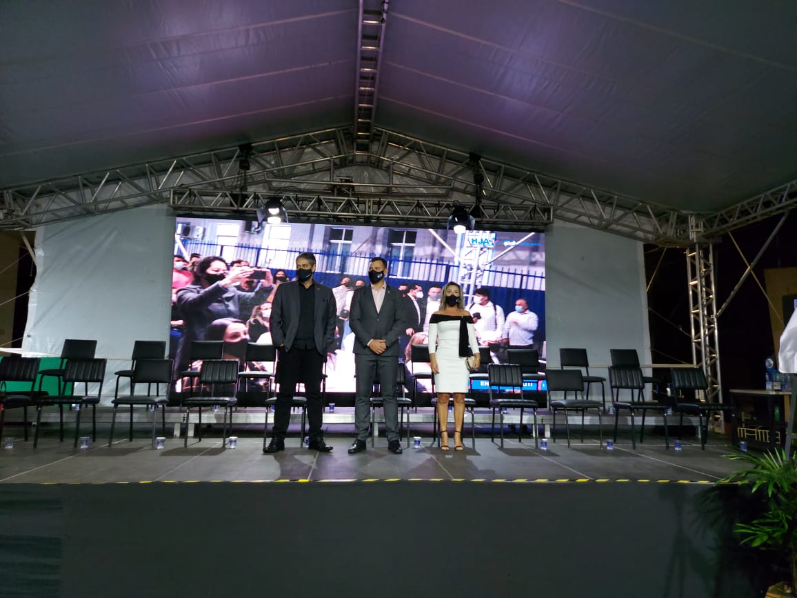 Três pessoas, duas de terno escuro e uma mulher de vestido branco, estão em pé sobre um palco coberto, todas usando máscara. Ao fundo, há um telão grande exibindo a imagem de uma plateia e outras pessoas. Cadeiras vazias estão dispostas no palco, sugerindo um evento formal ou cerimônia.