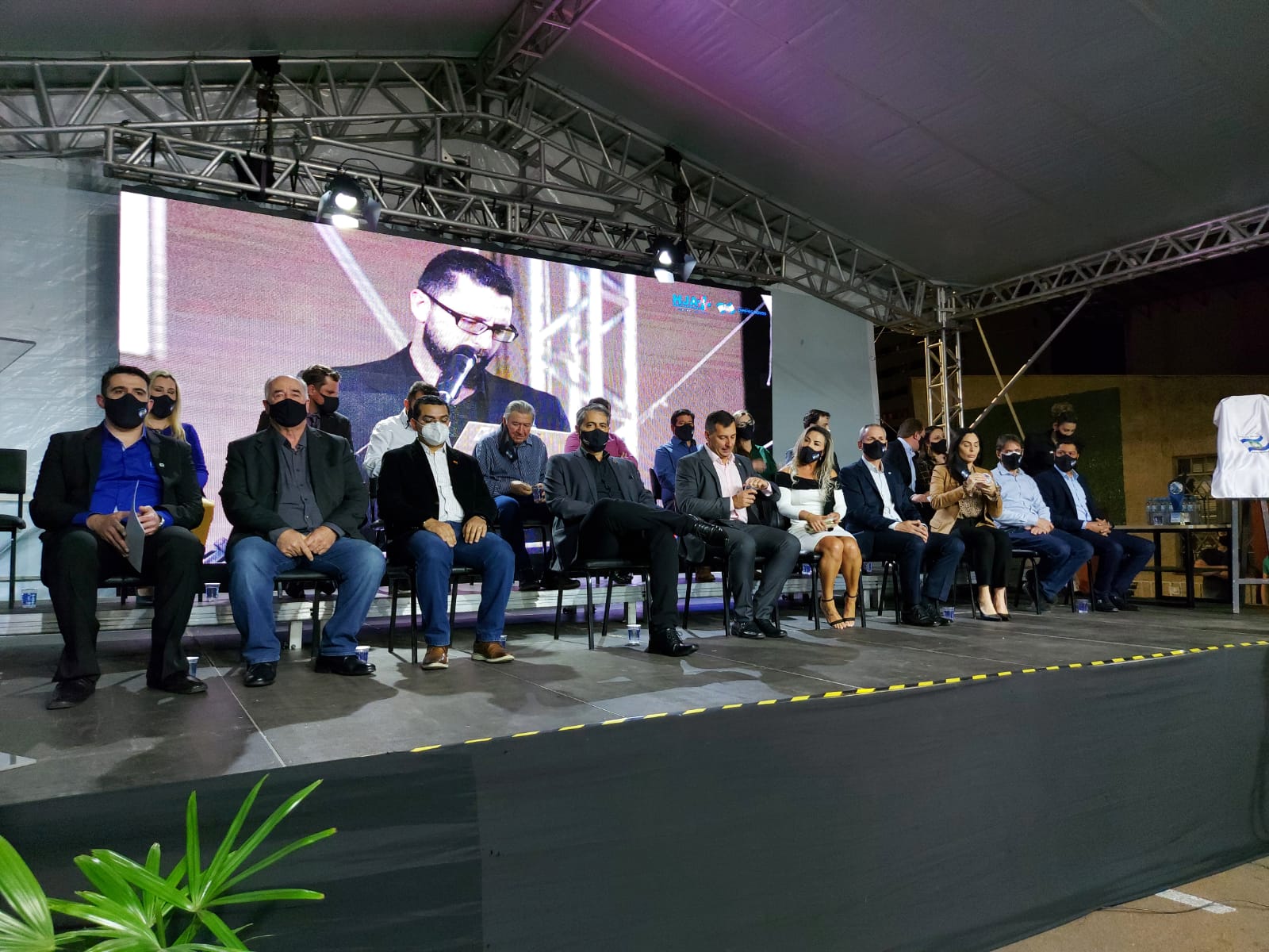 Grupo de pessoas sentadas em duas fileiras sobre um palco coberto, todas usando máscaras. Ao fundo, um telão exibe a imagem ampliada de um homem falando ao microfone. O ambiente sugere um evento formal ou cerimônia, com iluminação artificial e estrutura metálica.