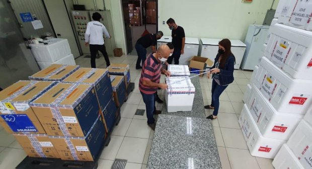 Sala com várias caixas térmicas brancas empilhadas e caixas de papelão organizadas. Algumas pessoas, usando máscaras, manuseiam as caixas sobre uma bancada de granito, sugerindo um ambiente de armazenamento ou distribuição de vacinas ou insumos de saúde.