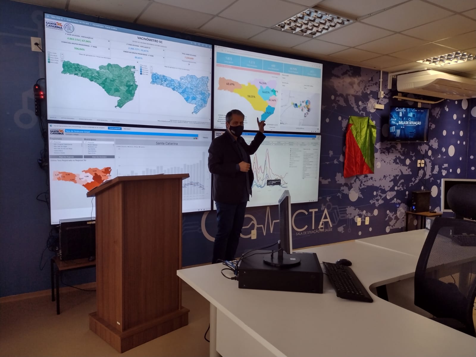 Homem de terno escuro e máscara está em pé ao lado de um púlpito, apontando para grandes telas na parede que exibem mapas e gráficos de dados de vacinação em Santa Catarina. O ambiente é uma sala de monitoramento, com mesas, computadores e decoração temática de saúde pública.