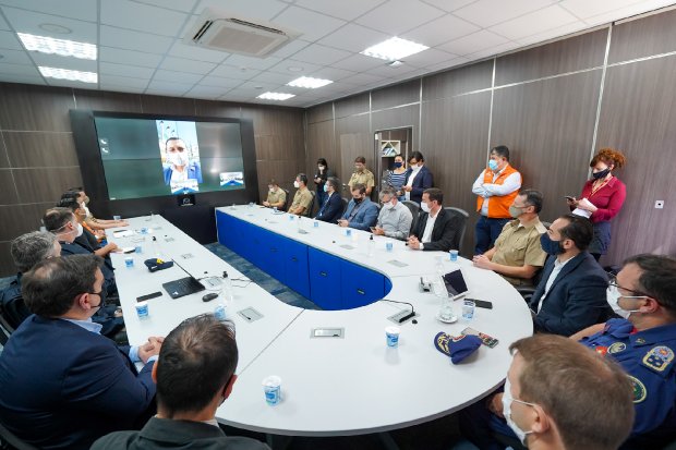 Sala de reuniões com mesa oval branca, onde várias pessoas, incluindo autoridades e profissionais de segurança, estão sentadas e algumas em pé, todas usando máscaras. Na parede ao fundo, uma grande tela exibe uma videoconferência com um homem participando remotamente. O ambiente é moderno e bem iluminado.