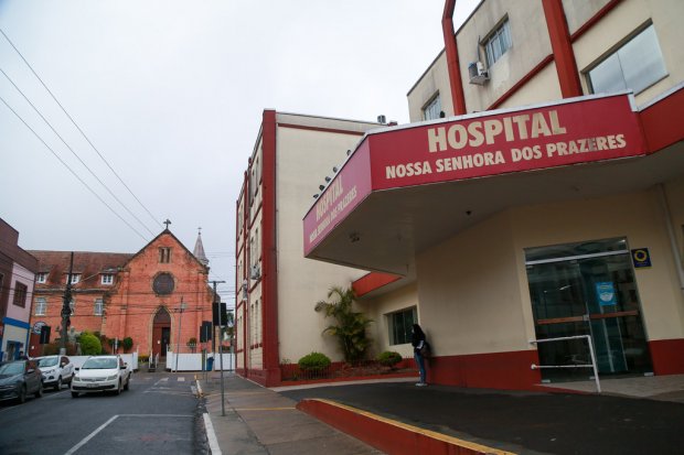 Fachada do Hospital Nossa Senhora dos Prazeres, com letreiro em destaque na entrada do prédio de cor bege e detalhes vermelhos. Ao fundo, do outro lado da rua, há uma igreja de tijolos aparentes. O cenário mostra uma rua urbana com alguns carros estacionados e uma pessoa próxima à entrada do hospital.
