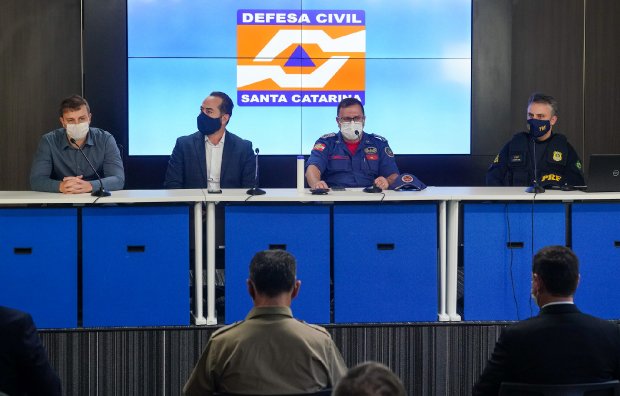 Quatro homens sentados atrás de uma mesa em uma coletiva de imprensa, todos usando máscaras. Ao fundo, um telão exibe o logotipo da Defesa Civil de Santa Catarina. Na frente da mesa, algumas pessoas assistem à apresentação, sugerindo um evento oficial ou reunião de autoridades.