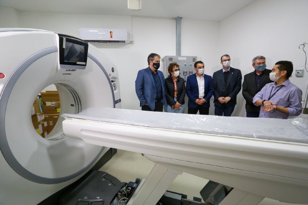 Grupo de seis pessoas usando máscaras observa um equipamento de tomografia computadorizada em uma sala branca de hospital. O aparelho está em destaque à esquerda, com a maca posicionada à frente, sugerindo uma visita técnica ou inauguração de setor de diagnóstico por imagem.