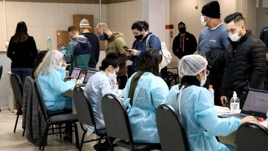Profissionais de saúde usando aventais, toucas e máscaras atendem pessoas em uma sala, sentados em mesas com computadores e frascos de álcool em gel. Várias pessoas, também de máscara, aguardam em pé para serem atendidas, sugerindo um ambiente de vacinação ou triagem em saúde.