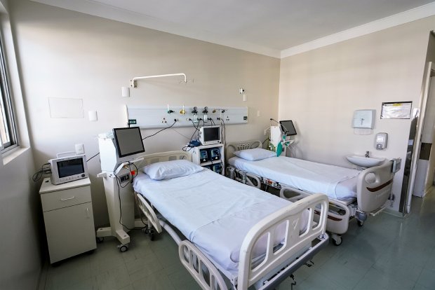 Quarto hospitalar com duas camas equipadas com lençóis brancos, monitores e aparelhos médicos ao lado de cada leito. O ambiente é limpo, iluminado por janela, e há uma pia com dispensador de sabonete fixado na parede.
