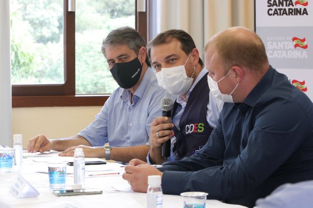 Governador Carlos Moisés sentado em mesa de reunião segurando microfone, acompanhado por duas autoridades (uma à esquerda e outra à direita), durante anúncio da ativação de 30 novos leitos de UTI na região Norte de Santa Catarina.