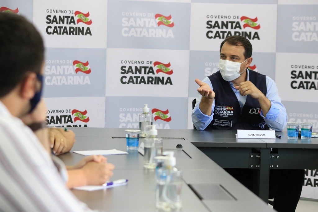 Em Rio Negrinho, governador responde perguntas, ele está sentado, de máscara com um colete azul escuro.