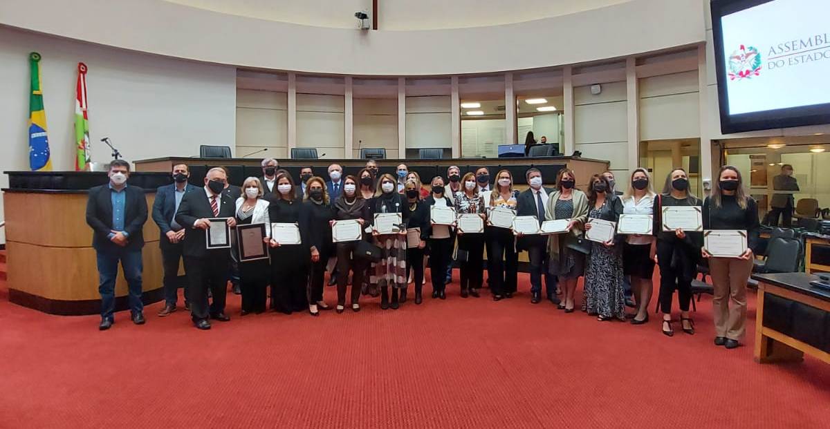 Grupo de cerca de 30 pessoas posa para foto em plenário da Assembleia Legislativa de Santa Catarina. Todos usam máscaras de proteção facial e seguram certificados. Ao fundo, há as bandeiras do Brasil e de Santa Catarina, e no canto direito, um telão exibe o logotipo da Assembleia Legislativa do Estado.