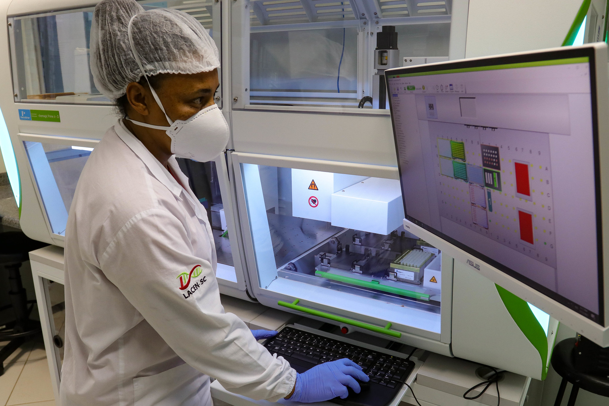 Profissional de laboratório com máscara, touca e luvas opera equipamento automatizado, acompanhando os resultados em um monitor de computador. No jaleco, está estampado o logotipo do LACEN/SC.