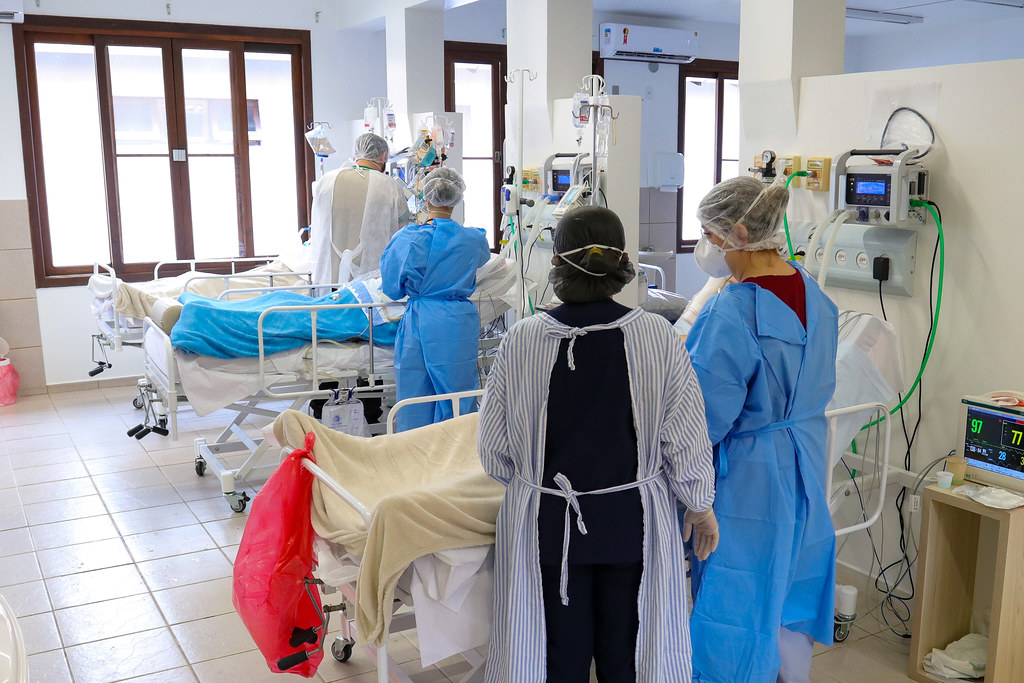 Enfermaria hospitalar com três pacientes internados, cada um acompanhado por um profissional de saúde em pé ao lado do leito, demonstrando a estrutura de 30 novos leitos de UTI ativados na região Norte de Santa Catarina.