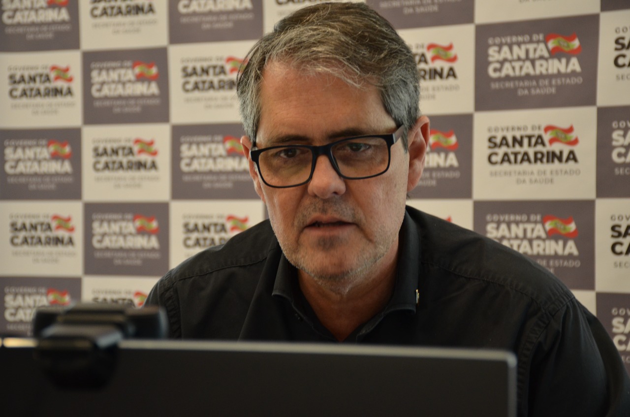 Homem de óculos e camisa preta participa de uma videoconferência ou entrevista, com expressão séria. Ao fundo, painel institucional com logotipos do eesa:'Governo de Santa Catarina - Secretaria de Estado da Saúde', repetidos em padrão quadriculado. Um notebook e uma webcam aparecem parcialmente em primeiro plano. 