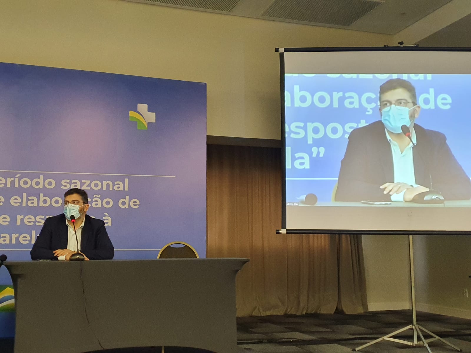 Homem com máscara cirúrgica fala ao microfone em uma mesa de conferência, com fundo azul exibindo parte do texto 'período sazonal de elaboração de resposta'. À direita, a mesma imagem é projetada em uma tela grande, mostrando o homem com a mesma postura e microfone à frente. O ambiente é fechado, com iluminação artificial e cortinas ao fundo.