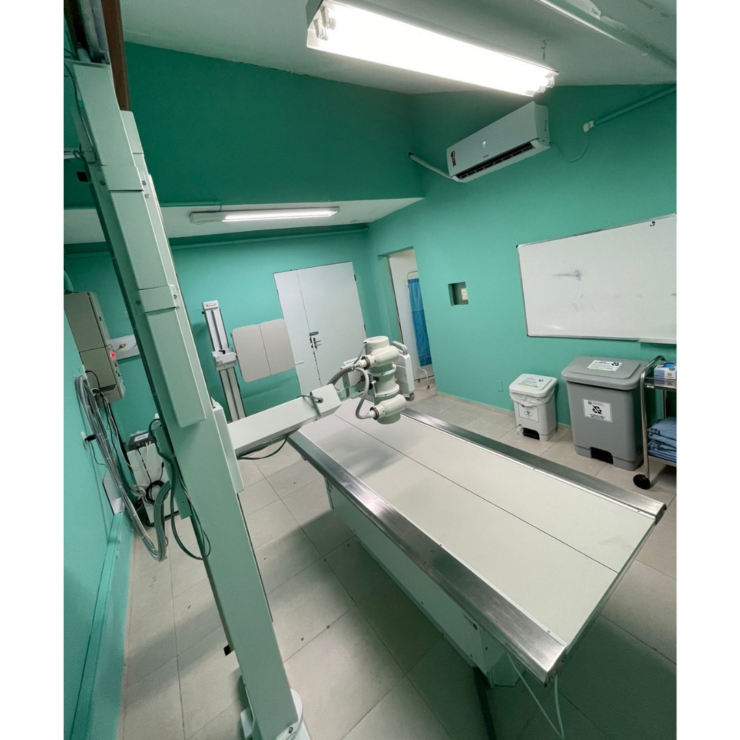 Sala de exame radiológico hospitalar com paredes pintadas de verde água. No centro da sala, há uma mesa de raio-X com superfície metálica e um equipamento radiológico montado acima dela, suspenso por um braço mecânico. À esquerda, há painéis e cabos conectados ao equipamento. Ao fundo, vê-se uma porta dupla, um suporte vertical de exames e uma cortina azul parcialmente visível em uma sala adjacente. À direita, há um quadro branco, lixeiras para descarte de resíduos hospitalares com símbolos de material infectante e reciclável, e uma prateleira com lençóis azuis dobrados. Um aparelho de ar-condicionado está instalado na parede superior. A sala é bem iluminada por lâmpadas fluorescentes no teto.