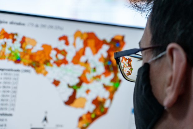 Pessoa usando máscara preta e óculos observa atentamente a tela de um computador que exibe um mapa colorido do estado de Santa Catarina. O mapa apresenta diferentes regiões em tons de laranja, branco e verde, indicando dados estatísticos, possivelmente relacionados à saúde ou à pandemia. A imagem reflete o mapa nas lentes dos óculos da pessoa, destacando o foco na análise geográfica. A atmosfera sugere um ambiente técnico ou de monitoramento de dados.