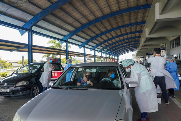 Profissionais de saúde aplicam vacinas em um sistema drive-thru sob uma cobertura metálica azul. Vários carros estão parados em fila, com pessoas sendo atendidas dentro dos veículos. Todos os profissionais utilizam equipamentos de proteção individual, como aventais, toucas, luvas e máscaras. A cena transmite um esforço organizado de vacinação em massa.
