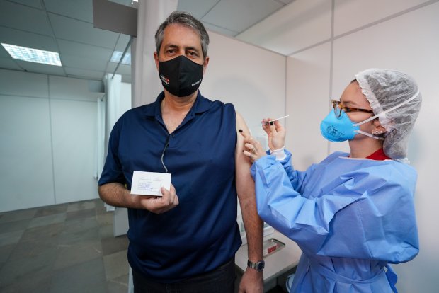 Homem de máscara preta recebe uma dose de vacina no braço esquerdo, aplicada por uma profissional de saúde com máscara azul, touca e avental de proteção. Ele segura um cartão de vacinação com a mão direita e olha para a câmera, enquanto está em pé dentro de um ambiente clínico com paredes brancas.