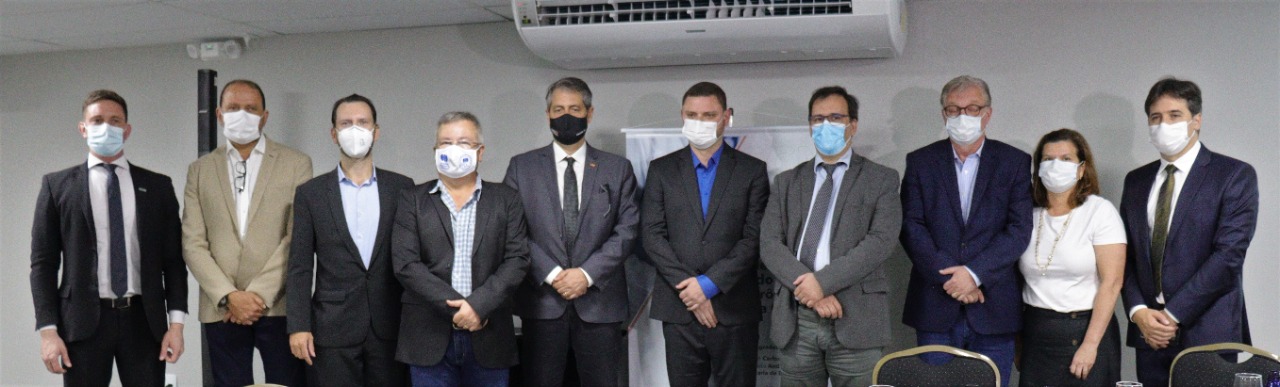 Grupo de onze pessoas, todos usando máscaras, posam lado a lado para uma foto em ambiente interno, durante um evento ou reunião. A maioria veste trajes formais, como ternos e blazers. Ao fundo, há um aparelho de ar-condicionado e parte de um banner institucional parcialmente visível. Homens e uma mulher compõem o grupo, demonstrando um encontro profissional.