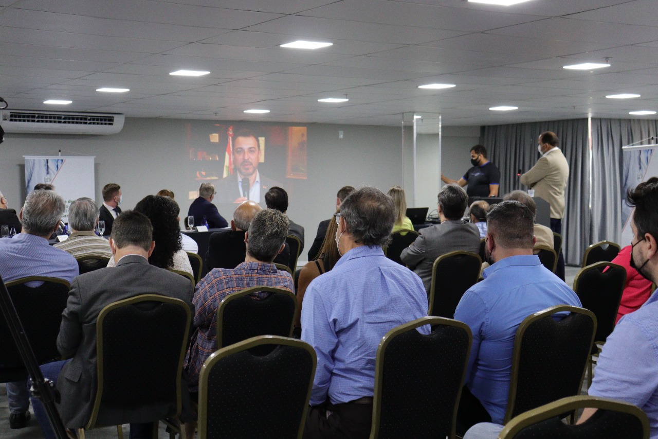 Auditório com várias pessoas sentadas assistindo a uma videoconferência projetada em uma parede. A imagem na projeção mostra um homem falando, em cenário formal. A maioria dos presentes está de máscara e veste roupas sociais. No canto direito da imagem, duas pessoas estão em pé, uma operando equipamentos e outra ao lado de um púlpito, sugerindo uma participação híbrida entre presencial e remota em um evento corporativo.