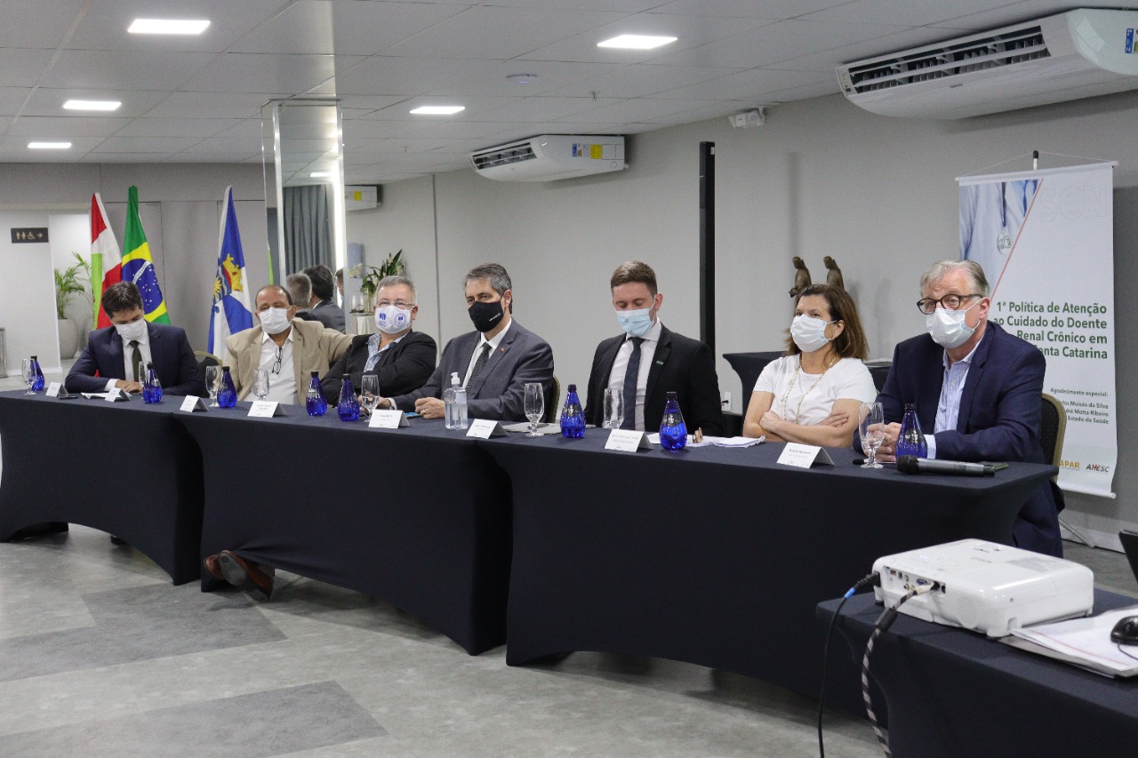 Autoridades e profissionais sentados em uma mesa de reunião formal, todos usando máscaras. São sete pessoas, incluindo uma mulher, participando da discussão sobre a 1ª Política de Atenção ao Cuidado do Doente Renal Crônico em Santa Catarina, conforme indicado no banner ao fundo. Bandeiras do Brasil, de Santa Catarina e de outra entidade estão dispostas ao fundo. Sobre a mesa, há garrafas de água azuis, taças e crachás de identificação. Um projetor está posicionado em primeiro plano, sugerindo apoio audiovisual.