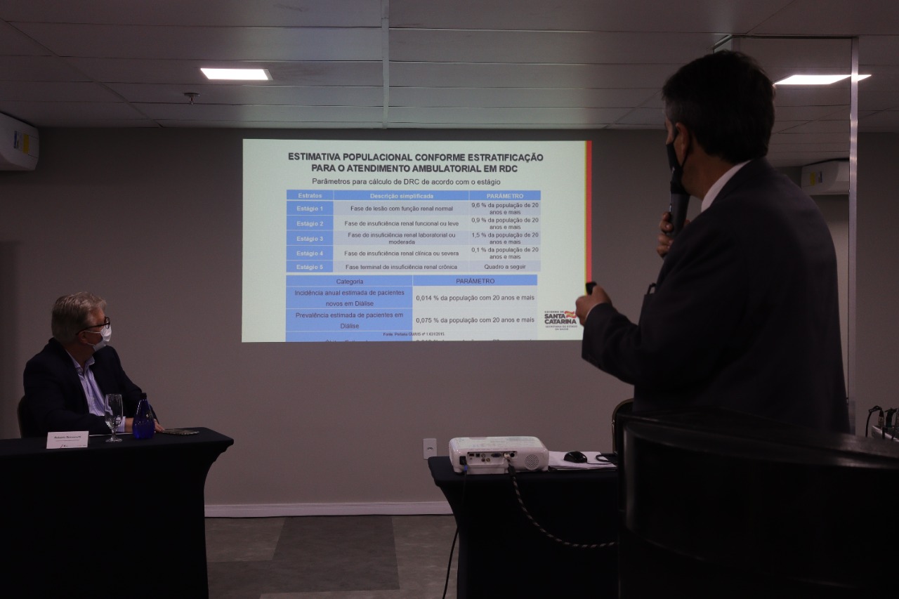 'Homem de terno e máscara preta faz uma apresentação utilizando microfone e apontador, enquanto projeta uma tabela com o título 'Estimativa populacional conforme estratificação para o atendimento ambulatorial em RDC'. A tabela exibe estágios da doença renal crônica e os respectivos parâmetros populacionais. À esquerda, outro homem de máscara, vestido com blazer azul, acompanha a apresentação sentado à mesa. Na frente, há um projetor sobre a mesa com fios conectados.