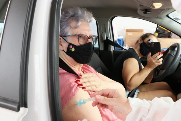Senhora de blusa rosa recebendo vacina contra COVID-19 dentro do carro em posto drive-thru, enquanto passageira ao lado registra o momento com celular preto - demonstrando a aceleração da campanha em Santa Catarina.