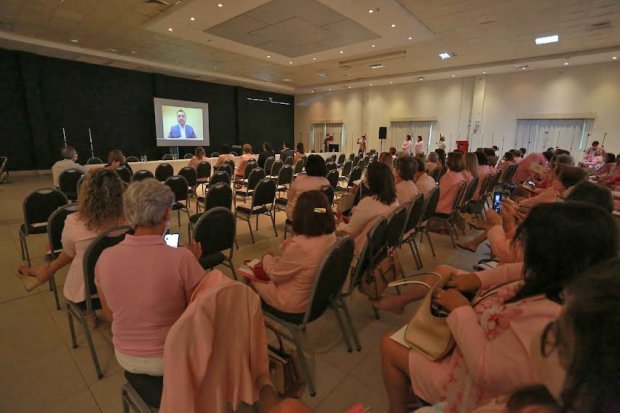 Público sentado em um auditório assiste à projeção de um vídeo com um homem falando. A maioria das pessoas presentes veste roupas em tons de rosa, sugerindo um evento relacionado à saúde da mulher, como uma campanha do Outubro Rosa. O local é amplo, com iluminação de teto e cadeiras organizadas em fileiras. Algumas participantes estão usando celular, enquanto outras acompanham atentamente a apresentação.