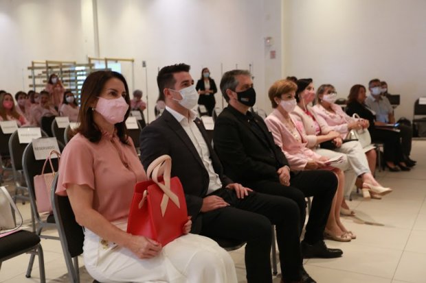 Pessoas sentadas nas primeiras fileiras de um auditório participam atentamente de um evento. Em destaque, uma mulher de máscara rosa segura uma bolsa vermelha com um laço símbolo da campanha Outubro Rosa. Ao lado dela, dois homens de terno e outras mulheres com roupas em tons de rosa e branco também usam máscaras. Ao fundo, mais participantes, em sua maioria mulheres, estão sentadas em cadeiras organizadas, reforçando o clima institucional e de conscientização sobre a saúde feminina.