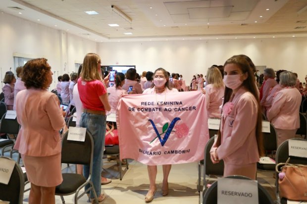 Mulheres reunidas em um auditório participam de evento da Rede Feminina de Combate ao Câncer. Em destaque, uma mulher ao centro segura uma bandeira rosa com o nome da entidade e o texto 'Balneário Camboriú'. Outras participantes, a maioria vestindo roupas cor-de-rosa e máscaras, estão em pé, interagindo e registrando o momento com celulares. Cadeiras com plaquinhas de 'RESERVADO' estão dispostas em fileiras, reforçando o clima organizado e solene do encontro.
