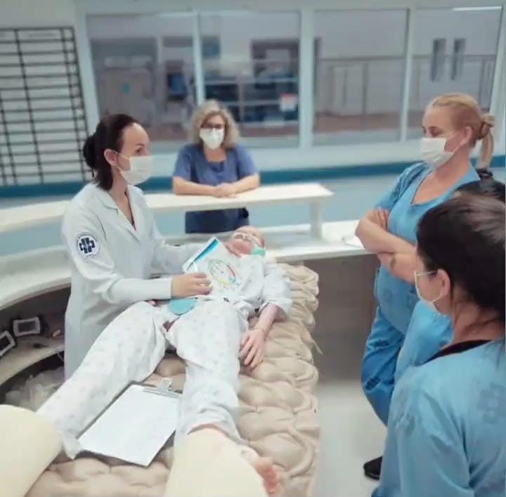 Profissionais de saúde em ambiente hospitalar participam de um treinamento prático. Quatro mulheres — três com roupas azul claro e uma com jaleco branco — estão reunidas em volta de um manequim médico simulado de paciente deitado em uma maca. A profissional de jaleco segura uma prancha anatômica e parece explicar um procedimento, enquanto as outras observam com atenção. Todas usam máscaras faciais. Ao fundo, outra mulher observa a cena de trás de um balcão.