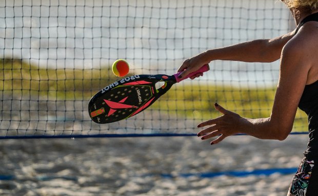 Mulher praticando tênis com raquete preta e rosa, rebatendo bola amarela e laranja à beira da lagoa em Santa Catarina.