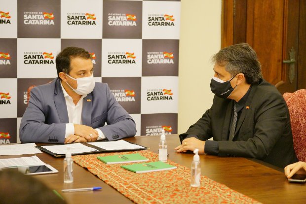 Governador Carlos Moisés em reunião com gestor de saúde em mesa de reuniões, com logotipos do Governo de Santa Catarina ao fundo.