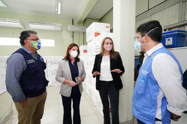 Nova secretária da Saúde e governadora de SC acompanhadas por dois técnicos em vistoria à central de distribuição de vacinas, com diversas caixas térmicas espalhadas pelo ambiente interno.