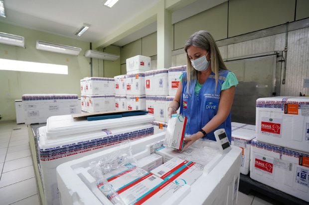 Profissional loira de casaco azul e máscara manipulando caixa térmica com doses de vacina em sala de distribuição repleta de outras caixas.