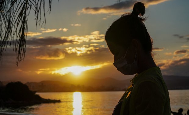 Perfil de mulher olhando para a esquerda com luz solar iluminando seu rosto, tendo praia e folhas de coqueiro ao fundo, simbolizando a continuidade das medidas de combate à COVID-19 em Santa Catarina durante o verão.