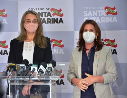 Secretária da Saúde de SC e assessora em trajes formais durante coletiva de imprensa, com parede de fundo contendo logotipos do Governo de Santa Catarina.