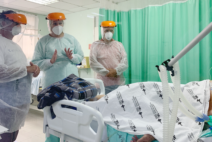 Três profissionais de saúde com EPIs completos (máscaras, protetores faciais e roupas de proteção) em pé ao lado do leito conversando com paciente internado no Hospital Regional de São Miguel do Oeste, durante o projeto 'Onde Há Vida, Há Esperança'.