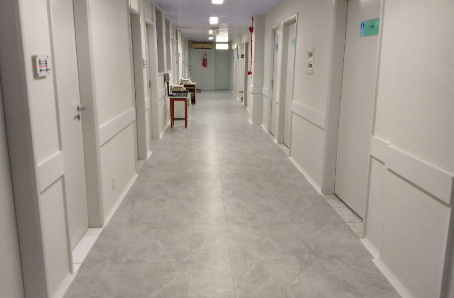 Corredor interno de hospital ou clínica, com piso de porcelanato cinza claro e paredes brancas. Diversas portas alinhadas dos dois lados, algumas com placas de identificação. Ao fundo, há uma mesa, cadeiras e um extintor de incêndio fixado na parede. O ambiente é limpo, organizado e bem iluminado por luzes de teto.