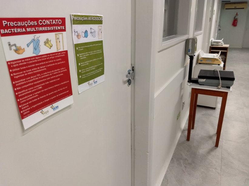 Corredor de hospital com cartazes afixados na parede indicando precauções de contato para bactéria multirresistente e precauções para aerossóis. Ao lado, há uma balança sobre uma mesa de madeira e outros equipamentos médicos. O ambiente é limpo, com piso claro e portas alinhadas.