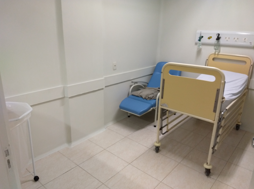 Quarto hospitalar simples com uma cama de paciente de grades amarelas, poltrona azul ao lado com um cobertor dobrado e parede branca. Há também um suporte para lixo no canto esquerdo e equipamentos médicos fixados na parede acima da cama. O ambiente é limpo e organizado.