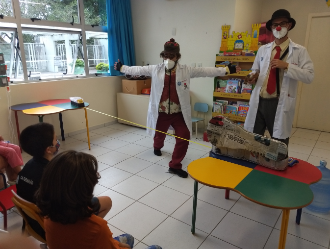 Dois palhaços de jaleco branco, nariz vermelho e máscara fazem uma apresentação para crianças em uma sala colorida, com mesas e cadeiras pequenas. Um dos palhaços segura uma fita amarela esticada, enquanto o outro está ao lado de uma maleta coberta de jornal. Ao fundo, há uma estante de livros infantis e uma janela grande. As crianças assistem sentadas, usando máscaras.