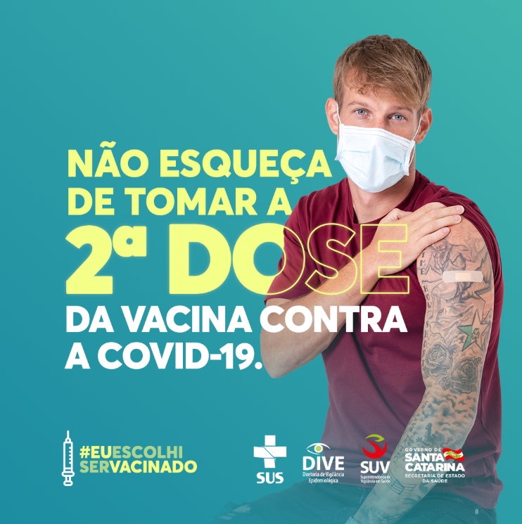 Banner com homem loiro de olhos azuis com máscara branca, roupa vermelha. Ele estende a manga curta de sua camisa mostrando seu ombro com um band-aid cobrindo onde foi aplicado vacina. No banner existe um texto: 'não esqueça de tomar a 2ª Dose da vacina contra a covid-19' 