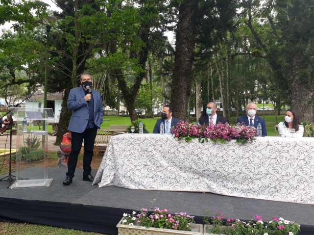 Homem de terno azul fala ao microfone em um palco ao ar livre, ao lado de uma mesa comprida coberta com toalha branca estampada e decorada com arranjos de flores roxas. À mesa, cinco pessoas estão sentadas, todas usando máscaras e trajes formais. O cenário é um jardim arborizado, com árvores altas ao fundo e ambiente de natureza ao redor. Um púlpito de acrílico com microfone e garrafa de água está à esquerda do orador.