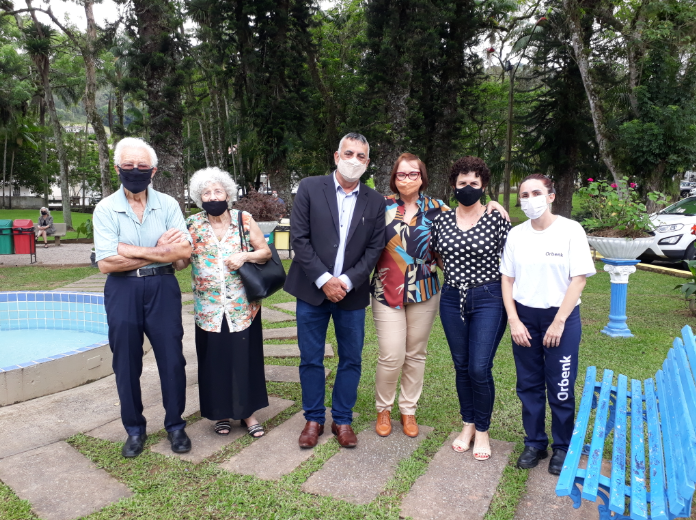 'Grupo de sete pessoas posa sorrindo para a foto em um jardim com árvores e flores ao fundo. Todos usam máscaras de proteção. À esquerda, um casal de idosos veste roupas casuais e está próximo a uma fonte azul clara. Ao centro, três pessoas usam roupas sociais, incluindo um homem de blazer escuro. À direita, duas mulheres usam roupas mais informais, sendo uma delas funcionária com uniforme da empresa 'Orbenk'. Ao fundo, há um banco azul, vasos de flores e carros estacionados.